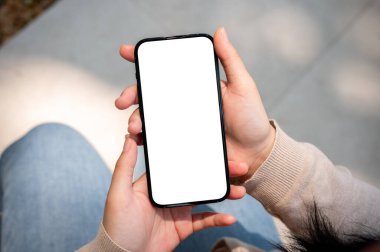 Güneşli bir günde kotlu bir kadının açık havada oturduğu ve akıllı telefonunu kullandığı yakın plan bir görüntü, beyaz ekran akıllı telefon modeli, bulanık bir arka planı olan bir kadının elinde..