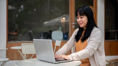 Konforlu giysiler içinde genç bir Asyalı kadın bir kafenin açık masasında çalışıyor, projesine odaklanıyor ya da bilgisayarındaki e-postalara cevap veriyor..