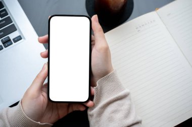 Modern bir ofis masasının üzerinde elinde beyaz ekran akıllı telefon olan bir kadının el resmi. İçinde bir kitap ve dizüstü bilgisayar var..