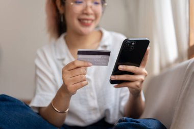 Genç, neşeli Asyalı bir kadın oturma odasında bir koltukta oturuyor, elinde kredi kartı ve akıllı bir telefon, mobil bankacılık uygulaması kullanıyor, evden alışveriş yapıyor ve faturaları ödüyor. Yakın çekim.