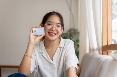 Genç mutlu bir Asyalı kadın oturma odasında bir koltukta oturuyor, kredi kartını gösteriyor ve kameraya gülümsüyor. nakit, bankacılık, yatırım, tasarruf, alışveriş, para