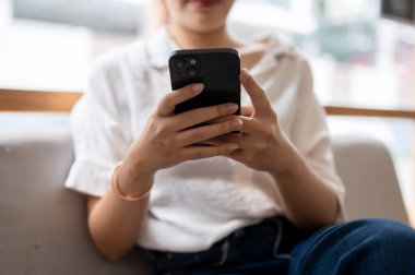 Oturma odasındaki kanepede oturmuş mesajları kontrol etmek ya da internet günlüğü okumak için akıllı telefonunu kullanan Asyalı bir kadının yakın plan görüntüsü. İnsanlar ve kablosuz teknoloji kavramları