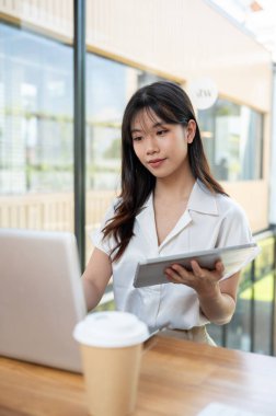 Çekici bir Asyalı iş kadını, dijital tablet ve dizüstü bilgisayarını kullanarak modern bir ofiste veya ortak çalışma alanında projesinde çalışıyor..