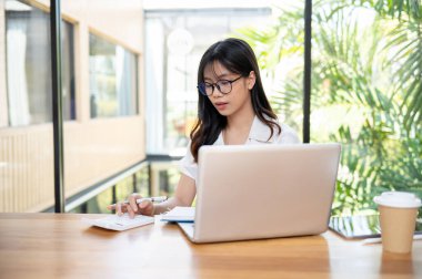 Güzel ve profesyonel bir Asyalı iş kadını ya da kadın muhasebeci hesap makinesi kullanıyor, iş harcamalarını hesaplamaya odaklanıyor, masadaki dizüstü bilgisayarla modern bir ofiste çalışıyor..