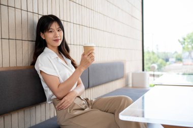 Çekici bir Asyalı kadın, elinde kahve fincanıyla çağdaş bir kahve dükkanında oturuyor ve kameraya gülümsüyor. Kahve molası, şehir hayatı, eğlence, insanlar ve yaşam tarzı konsepti