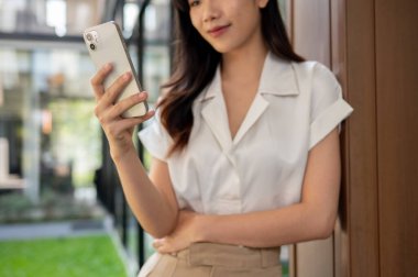 Milenyum Asyalı güzel bir iş kadınının bina duvarına yaslanıp akıllı telefonunu kullanmasının kırpılmış görüntüsü. İnternet günlükleri okumak, mesajları kontrol etmek, online videolar izlemek