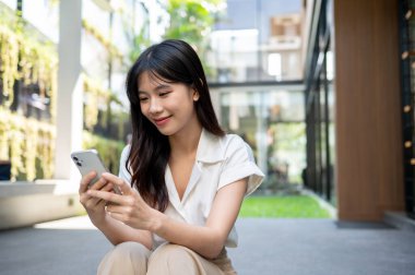 Güzel, mutlu bir Asyalı kadın binanın dışındaki merdivenlerde oturuyor ve akıllı telefonunu kullanıyor. sohbet, çevrimiçi blog okumak, online alışveriş, sosyal medyada videolar izlemek