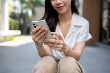 Güzel, mutlu bir Asyalı kadının binanın dışındaki merdivenlerde oturup akıllı telefonunu kullanışının yakın plan görüntüsü. sohbet, çevrimiçi blog okumak, online alışveriş, sosyal medyada videolar izlemek