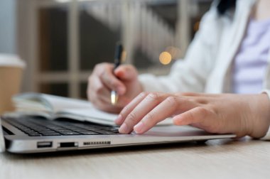 Kafede uzaktan çalışan, dizüstü bilgisayarını kullanan ve bir kitaba not alan bir kadının yakın plan fotoğrafı. Bilgisayarında ders çalışan ve bir kitaba not alan bir öğrenci..