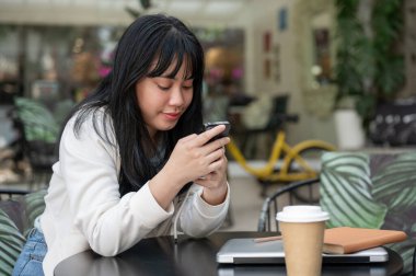 Genç, mutlu bir Asyalı kadın kafenin açık masasında otururken akıllı telefonunu kullanıyor. sohbet etmek, mesajları kontrol etmek, çevrimiçi bloglar okumak, sosyal medyada kaydırmak, online alışveriş