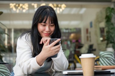 Genç, mutlu bir Asyalı kadın kafenin açık masasında otururken akıllı telefonunu kullanıyor. sohbet etmek, mesajları kontrol etmek, çevrimiçi bloglar okumak, sosyal medyada kaydırmak, online alışveriş