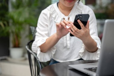 Asyalı bir kadının kesilmiş bir görüntüsü şehirdeki bir kafeden çalışıyor, açık bir masada oturuyor ve akıllı telefonunu kullanıyor. Metin okumak, sohbet etmek, mesajlara cevap vermek, mobil uygulama kullanmak