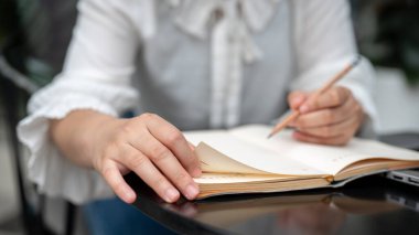 Masada oturan, elinde kalem tutan ve deftere yazı yazan bir kadının yakın plan görüntüsü. ödev yapmak, günlük tutmak, liste yapmak, yeni fikirler düşünmek.