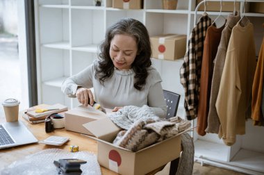 Tecrübeli bir Asyalı kadın terzi ya da terzi stüdyosundaki masasında makasıyla birlikte bir karton kutuyu açıyor..