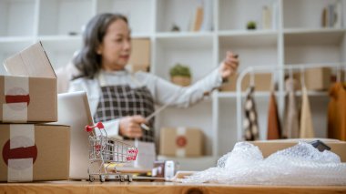 Bir alışveriş arabası modelinin seçici odak noktası, karton kutular, baloncuk ambalajı, bilgisayar, ve stüdyoda çalışan olgun Asyalı bir terzinin bulanık arka planına sahip bir masanın üzerindeki ekipman..