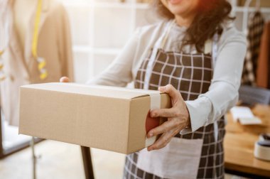 Yetişkin bir Asyalı kadın çevrimiçi dükkan satıcısının kırpılmış bir görüntüsü ev ofisinde dururken elinde bir karton kutu tutuyor. E-ticaret, çevrimiçi iş, girişimci, küçük işletme
