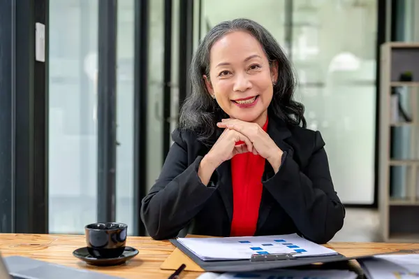 stock image A confident and attractive mature Asian businesswoman smiling at the camera while sitting at her desk in the office. female boss, manager, CEO, senior board member