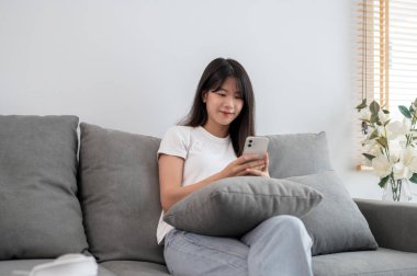 Rahat giysiler içinde güzel, pozitif, genç bir Asyalı kadın hafta sonu evdeki oturma odasında bir kanepede dinlenirken telefonuna kayıyor. insanlar ve yaşam tarzı konsepti