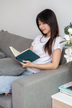 Genç, rahat bir Asyalı kadın evdeki oturma odasındaki kanepede kitap okuyor. ev hayatı, boş zaman ve yaşam tarzı