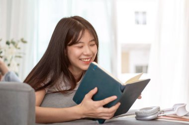 Güzel, mutlu, genç bir Asyalı kadın kanepede uzanıyor, kitap okumaktan ve oturma odasında dinlenmekten zevk alıyor. insanlar ve ev hayatı kavramları