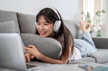 Kulaklık takan genç ve çekici bir Asyalı kadın oturma odasındaki kanepeye uzanıyor ve dizüstü bilgisayarını kullanıyor. İnternette sörf yapmak, sosyal medyada arkadaşlarla sohbet etmek.