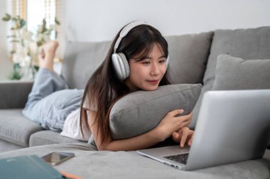 Kulaklık takan genç ve çekici bir Asyalı kadın oturma odasındaki kanepeye uzanıyor ve dizüstü bilgisayarını kullanıyor. İnternette sörf yapmak, sosyal medyada arkadaşlarla sohbet etmek.