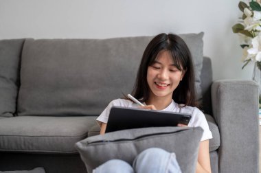 Dijital tabletinde stilus kalem kullanan mutlu bir Asyalı kadın oturma odasında dijital tabletini kullanıyor. Liste yapmak, çizmek, not almak, bir şeyler yazmak.