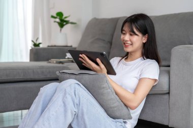 Genç, güzel bir Asyalı kadın hafta sonu evdeki oturma odasında dinlenirken dijital tabletini kullanıyor. Oyun oynamak, film izlemek, internette sörf yapmak, online blog okumak.