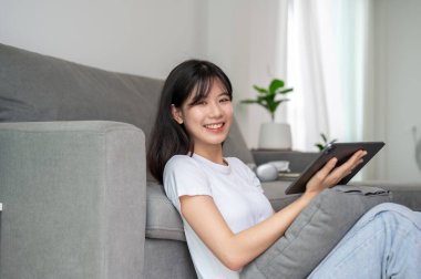 Rahat giysiler içinde çekici bir Asyalı kadın elinde dijital tabletle oturma odasında yere oturuyor ve kameraya gülümsüyor. İnsanlar, ev hayatı ve kablosuz teknoloji