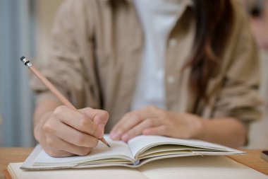 Kalem tutan, kapalı bir masadaki deftere bir şeyler yazan rahat elbiseli bir üniversite öğrencisinin yakın plan fotoğrafı. ödev yapmak, günlük tutmak, liste yapmak