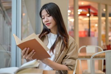 Genç, güzel, pozitif Asyalı bir kadın gündelik giysiler içinde alışveriş merkezindeki modern bir kafede kitap okuyor. insanlar ve şehir hayatı kavramları
