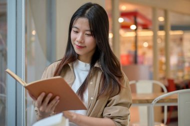 Genç, güzel, pozitif Asyalı bir kadın gündelik giysiler içinde alışveriş merkezindeki modern bir kafede kitap okuyor. insanlar ve şehir hayatı kavramları
