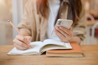 Kafedeki bir masada deftere bir şey yazarken akıllı telefonunu tutan bir kadının kırpılmış görüntüsü. ödev yapmak, not almak, liste yapmak, ders yazmak.