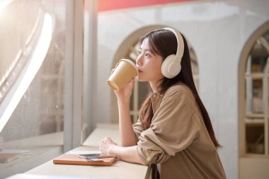 Güzel, rahat genç bir Asyalı kadın bir kafede kahve yudumlarken ve kulaklıklarıyla müzik dinlerken dışarıdaki manzarayı seyrediyor ve hayal kuruyor. Boş zaman, eğlence, mutluluk.