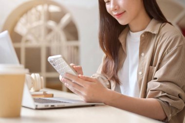 Günlük giysiler içinde güzel, mutlu bir Asyalı kadının kesilmiş bir görüntüsü bir kahve dükkanında dizüstü bilgisayarıyla oturuyor ve akıllı telefonunu kullanıyor. Uzaktan çalışan, üniversite öğrencisi, dijital göçebe.