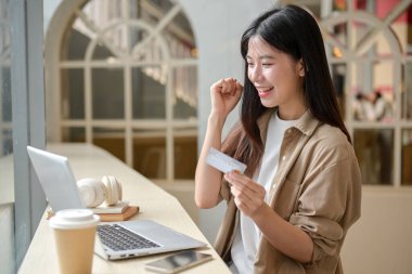 Genç ve güzel bir Asyalı kadın elinde kredi kartıyla mutlu bir ifadeyle yumruğunu sıkıp internetten alışveriş yapmaktan zevk alıyor. insanlar, yaşam tarzları ve finansal kavramlar