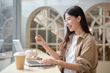 Bilgisayarından alışveriş yapan, elinde kredi kartı olan ve bir kafede bilgisayarını kullanan güzel bir Asyalı kadın. çevrimiçi ödeme, nakitsiz, mali yatırım