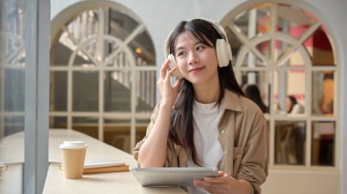 Genç ve güzel bir Asyalı kadın kulaklıklarıyla müzik dinliyor, dijital tabletiyle masada oturuyor ve kafenin dışındaki manzarayı seyrediyor..