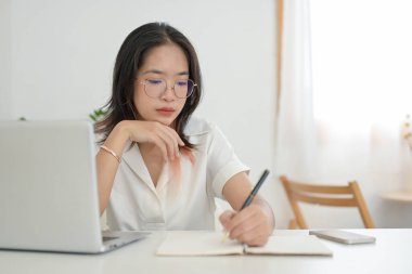 Genç odaklı Asyalı bir kadın bir kafeden uzaktan çalışıyor, not alıyor veya not defterine not alıyor, masada dizüstü bilgisayar var. Ödev yapan bir öğrenci ya da bir kafede internetten çalışan bir öğrenci.