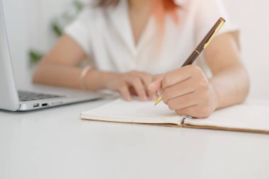 Asyalı bir kadının kalem tutarken, bir not defterine yazarken ya da not alırken bir kafeden uzaktan çalışırken çekilmiş görüntüsü. Ödev yapan ya da günlük tutan bir öğrenci,