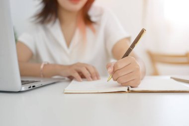 Asyalı bir kadının kalem tutarken, bir not defterine yazarken ya da not alırken bir kafeden uzaktan çalışırken çekilmiş görüntüsü. Ödev yapan ya da günlük tutan bir öğrenci,