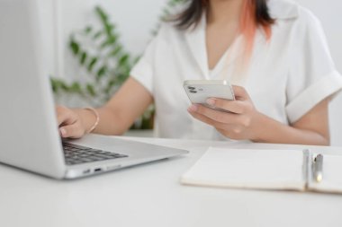 Kafede uzaktan çalışan Asyalı bir kadının kapalı bir masada dizüstü bilgisayarı üzerinde çalışırken akıllı telefonunu tutarken çekilmiş görüntüsü. İnsanlar ve kablosuz teknoloji kavramları