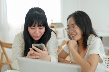 İki neşeli genç Asyalı kadın bir kafede oturmuş, akıllı telefon ekranına bakarken bir şeyler konuşuyorlar. insanlar ve yaşam tarzı kavramları