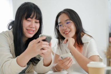 İki neşeli genç Asyalı kadın bir kafede oturmuş, akıllı telefon ekranına bakarken bir şeyler konuşuyorlar. insanlar ve yaşam tarzı kavramları