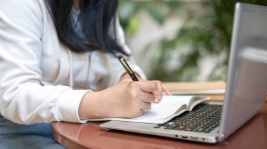 Asyalı bir kadının günlük kıyafetlerle çekilmiş yakın plan bir görüntüsü dizüstü bilgisayarında video izlerken ve kafede uzaktan çalışırken defterine bir şeyler yazıyor. Bir öğrenci çevrimiçi çalışıyor..