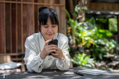 Parlak bir günde, arka bahçedeki ahşap bir masada akıllı telefonunu kullanan genç bir Asyalı kadın internet günlüklerini okuyor, mesajları cevaplıyor, sosyal medyada kaydediyordu..