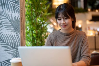 Genç ve çekici bir Asyalı kadın bir kafede çalışıyor, dizüstü bilgisayarı üzerinde çalışıyor. İnsanlar, modern hayat ve teknoloji kavramları