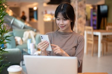 Genç ve çekici bir Asyalı kadın bir kafede dizüstü bilgisayarıyla otururken akıllı telefonunu kullanıyor, kafeden uzaktan çalışıyor. İnsanlar ve kablosuz teknoloji kavramları