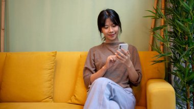 Çekici genç bir Asyalı kadın oturma odasındaki modern sarı bir kanepede akıllı telefonunu kullanıyor. Sohbet etmek, telefonda kaymak, mobil uygulama kullanmak, online alışveriş