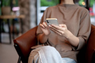 Pozitif Asyalı bir kadının kesilmiş bir görüntüsü modern bir kahve dükkanında akıllı telefonunu bir koltukta kullanıyor. sohbet, online alışveriş, mobil uygulama, sosyal medya, ağ kurma